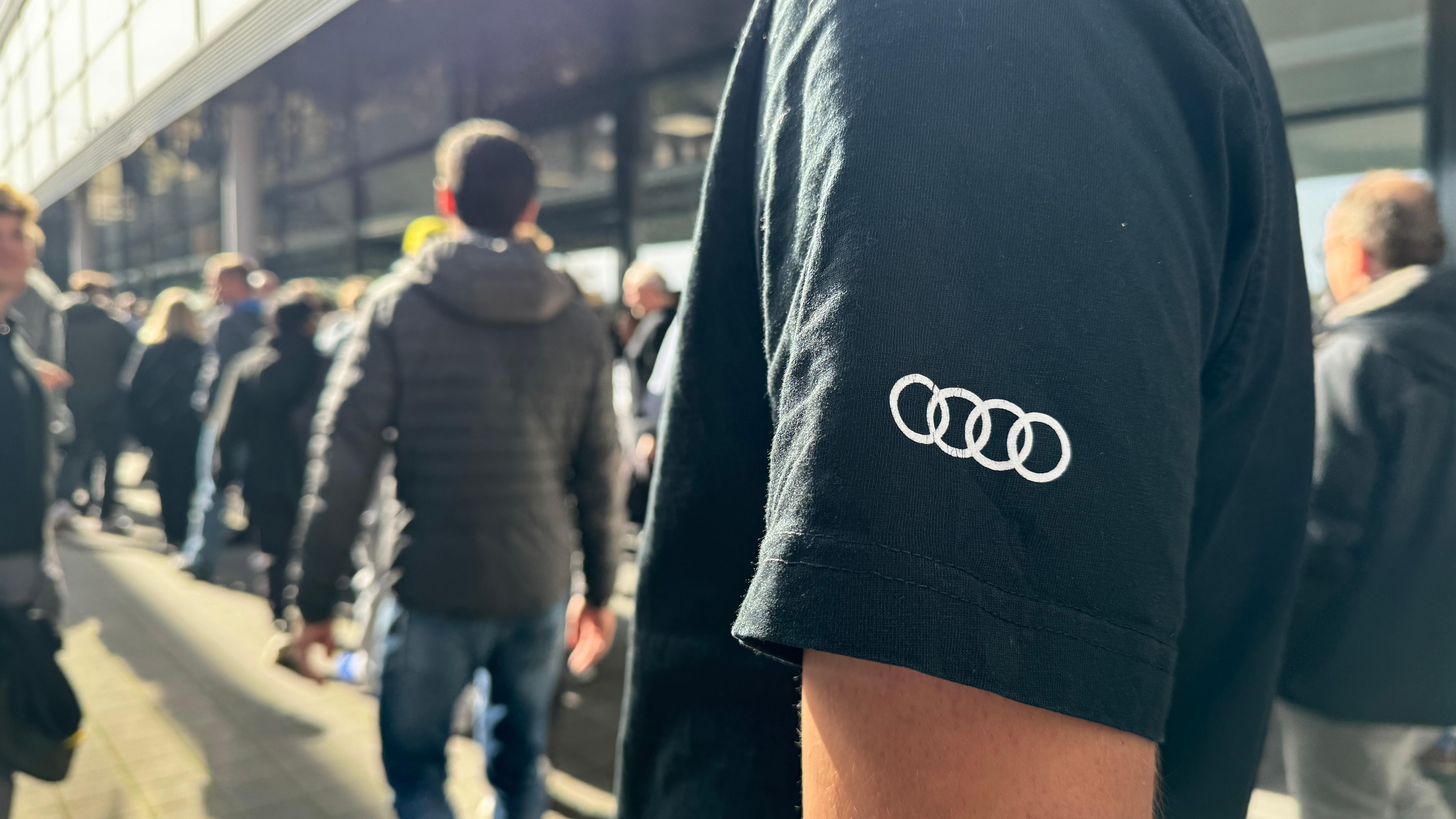 Im Vordergrund sieht man ein T-Shirt mit Audi-Logo. Dahinter stehen Menschen in einer Schlange vor der Halle, in der die Betriebsversammlung stattfindet.