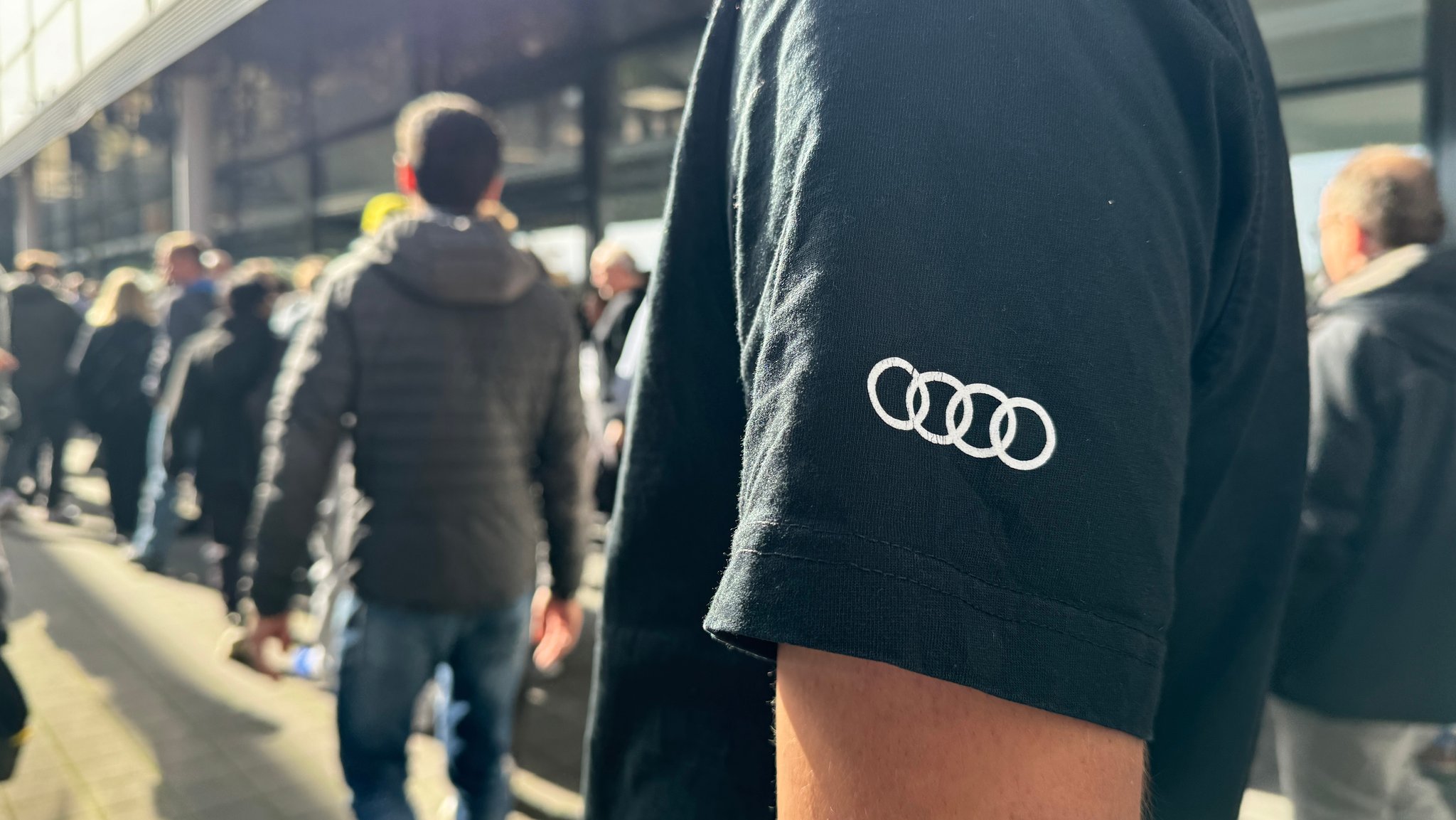 Im Vordergrund sieht man ein T-Shirt mit Audi-Logo. Dahinter stehen Menschen in einer Schlange vor der Halle, in der die Betriebsversammlung stattfindet.