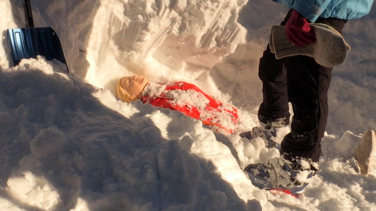Eine Puppe liegt tief verschüttet im Schnee – daneben die Lawinenschaufel, mit der sie ausgegraben wurde.