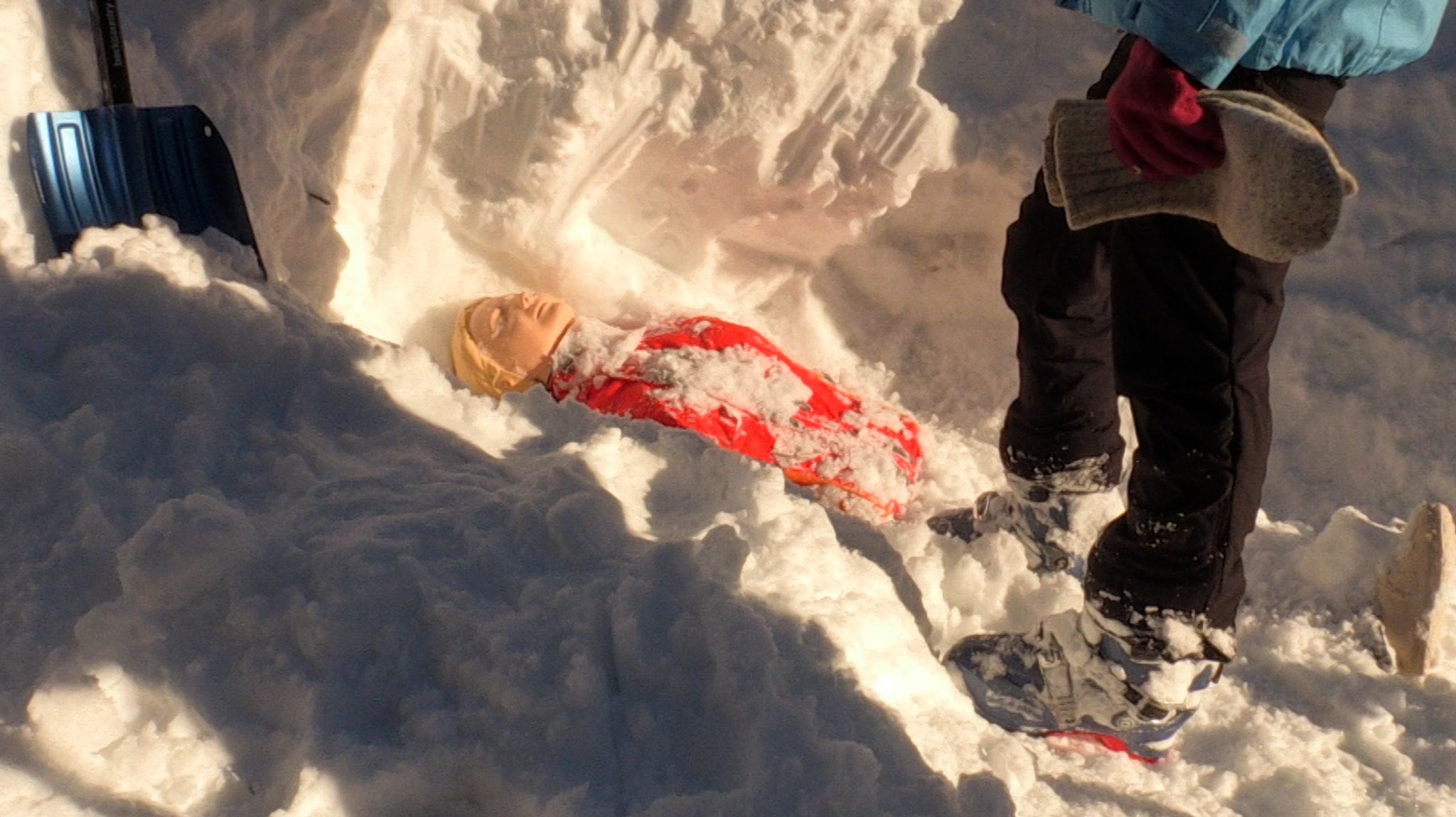 Eine Puppe liegt tief verschüttet im Schnee – daneben die Lawinenschaufel, mit der sie ausgegraben wurde.