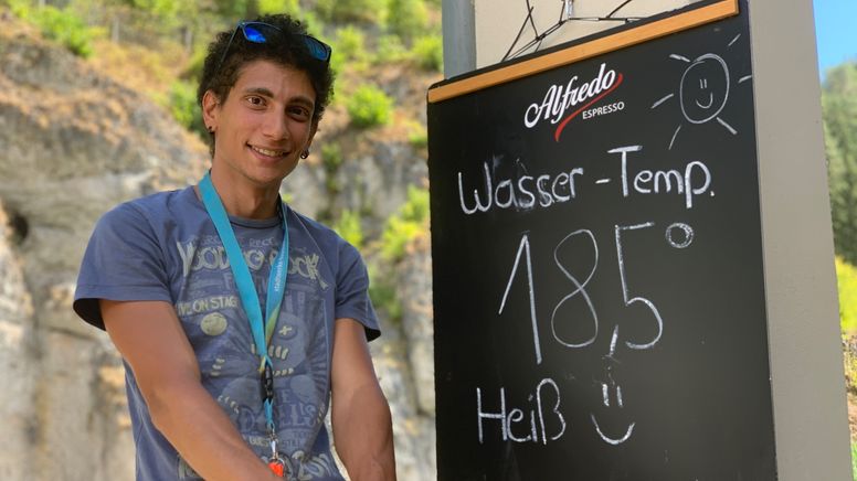Ein junger Mann steht neben einer Tafel mit der Aufschrift "Wasser-Temp. 18,5 Grad. Heiß"  | Bild:BR/Kristina Kreutzer