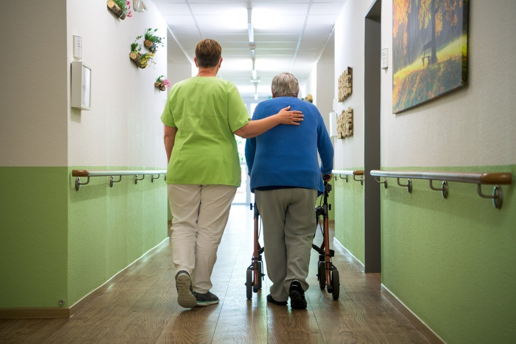 Eine Pflegerin geht gemeinsam mit dem Bewohner eines Seniorenheims den Gang entlang. (Symbolbild)