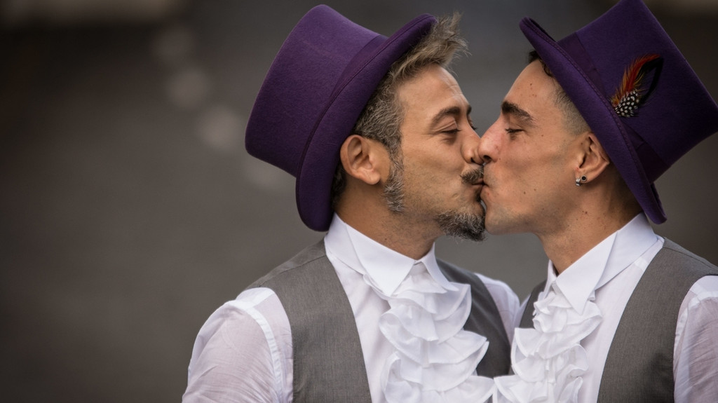 Zwei Männer in weißen Rüschenhemden, grauen Westen und violetten Zylinderhüten küssen sich. (Symbolbild)