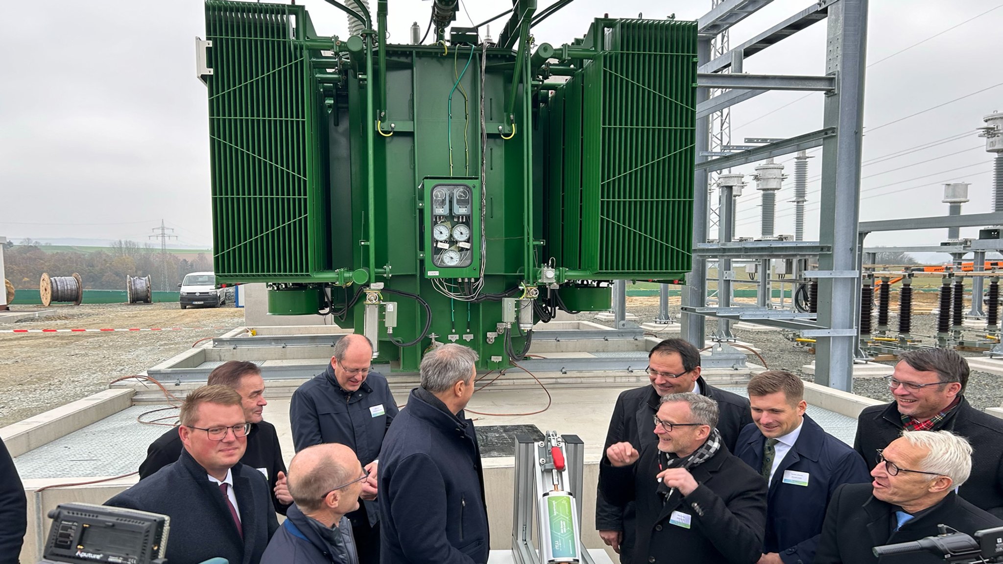 Geladene Gäste stehen vor einem großen grünen technischen Gerät bei der Einweihung des Batteriespeichers in Arzberg.