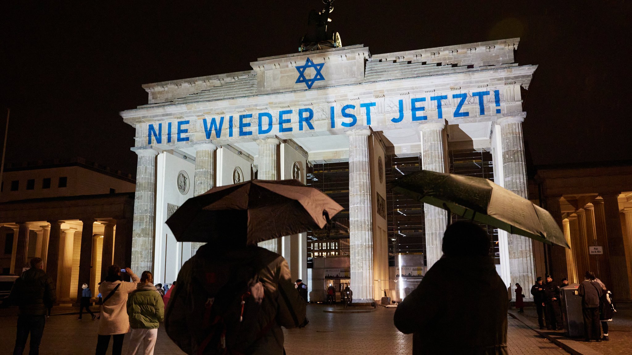 Auf das Brandenburger Tor ist in blau der Schriftzug "Nie wieder ist jetzt" projiziert, davor stehen Menschen mit Regenschirmen.