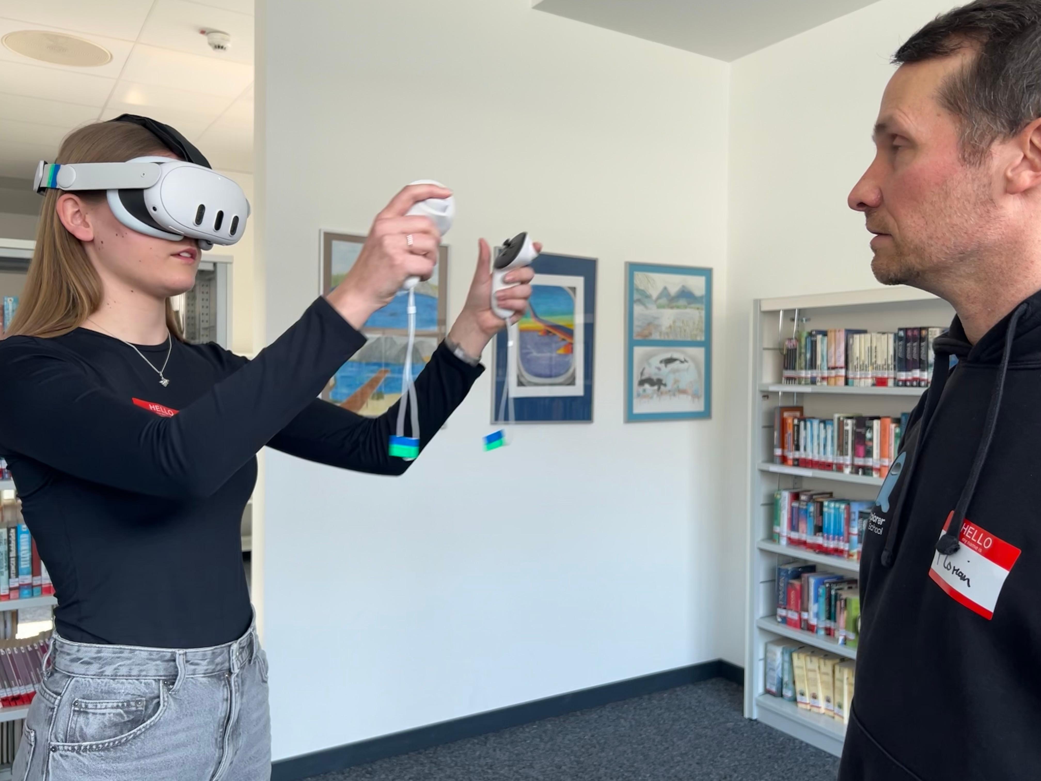 Eine Schülerin trägt eine VR-Brille und hat passende Controller in der Hand. Ein Mann sieht ihr dabei zu.