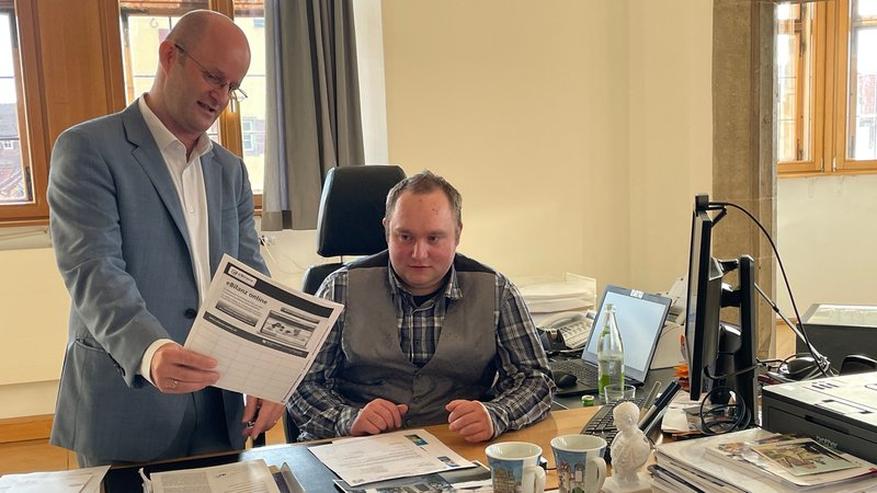 Zwei Männer sitzen an einem Schreibtisch und schauen ein Prospekt an. | Bild: BR / Julia Demel Zwei Männer sitzen an einem Schreibtisch und schauen ein Prospekt an.