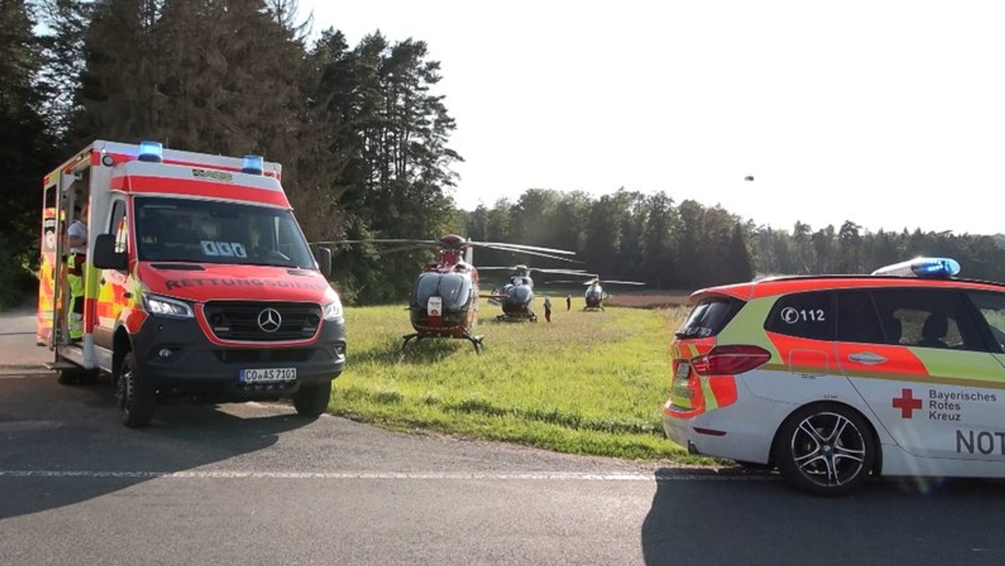 Auf einer Landstraße stehen zwei Rettungswagen, auf der Wiese daneben drei Hubschrauber, dazwischen bewegen sich Rettungskräfte.