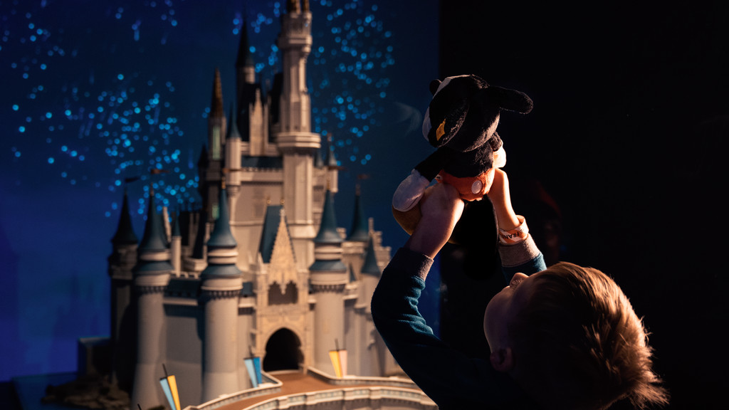 Ein Modell des Disney-Schlosses vor der Kulisse eines tiefblauen Nachthimmels, davor ein kleines Kind mit Micky Maus in den Händen