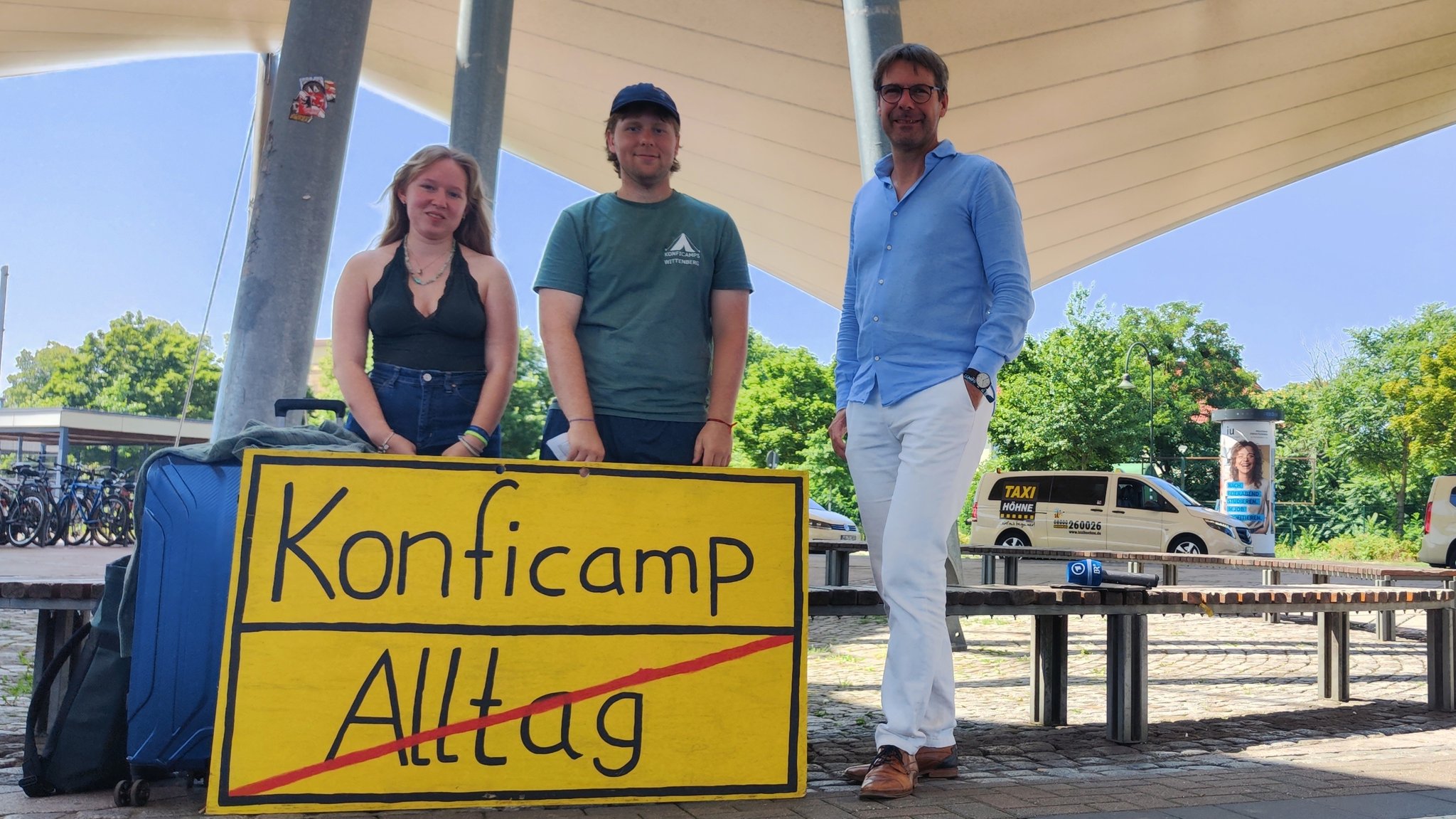 Pfarrer Maier steht mit zwei jungen freiwilligen Helfern vor einem gebastelten Ortsschild, auf dem "Konficamp" steht | Bild: Jasmin Brock/BR Pfarrer Maier steht mit zwei jungen freiwilligen Helfern vor einem gebastelten Ortsschild, auf dem "Konficamp" steht