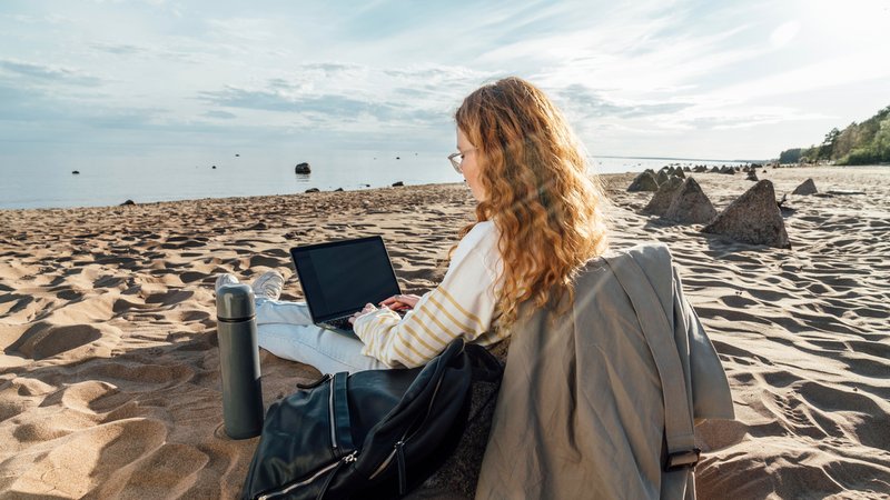 Eine junge Frau sitzt mit einem Laptop am Strand