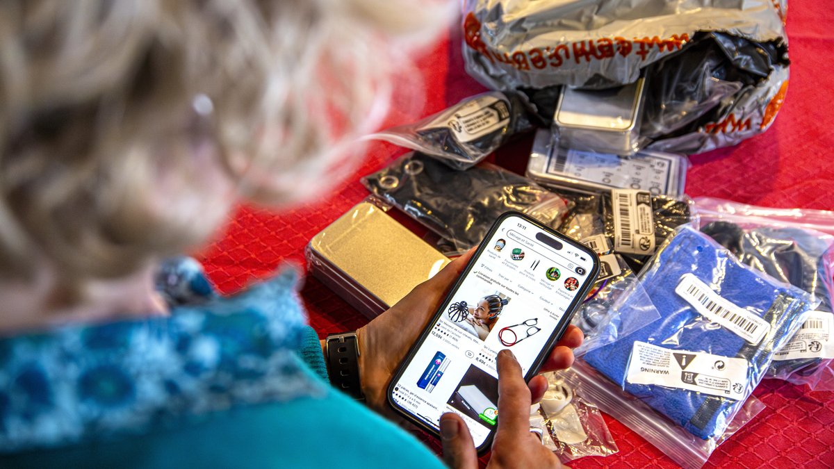 Viele in kleinen Plastiktüten verpacke Artikel liegen vor einer Versandtasche von Temu, im Vordergrund eine Person, die die Temu-App auf ihrem Handy geöffnet hat
