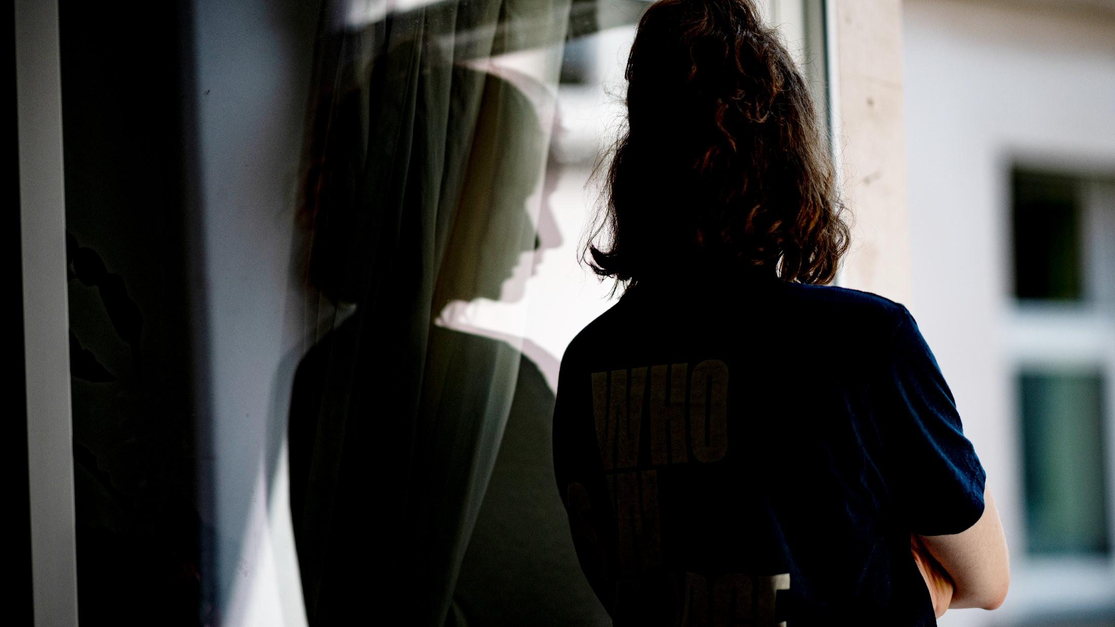 Eine Frau steht mit verschränkten Armen in ihrer Wohnung an einem Fenster.