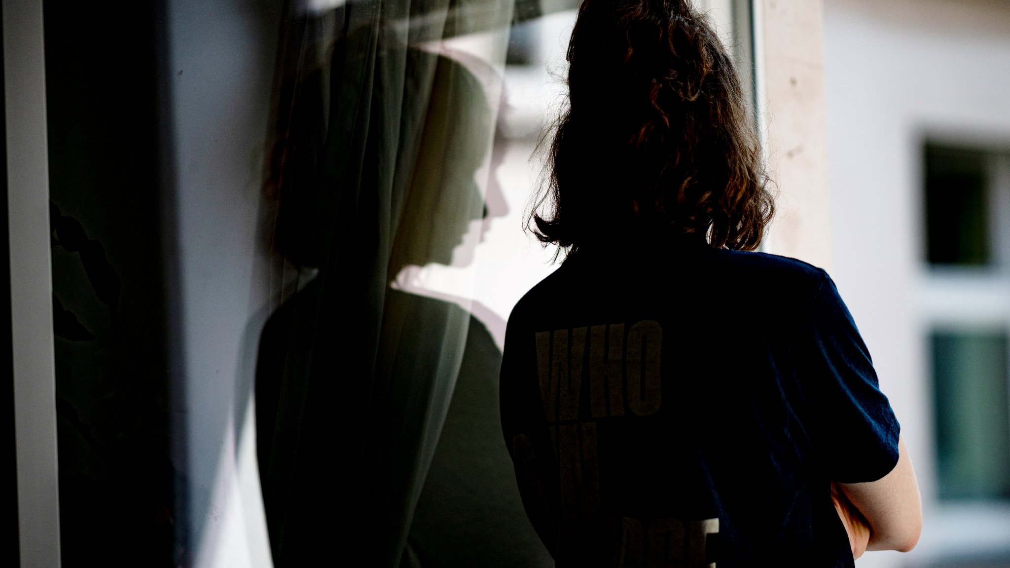 Eine Frau steht mit verschränkten Armen in ihrer Wohnung an einem Fenster.