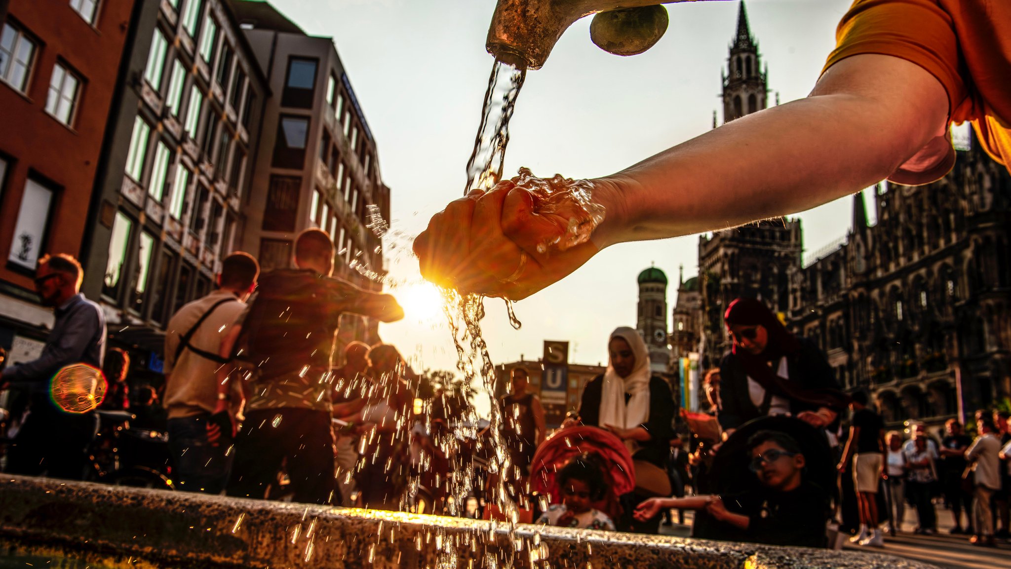 Sommerabend am Marienplatz: Brunnenwasser glitzert in der Abendsonne. Eine Frau läßt sich kaltes Wasser über die Hand laufen.