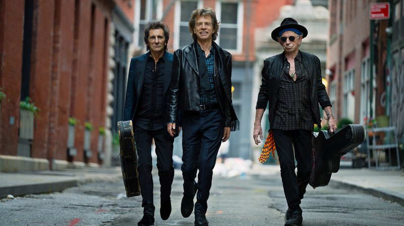 Ron Wood, Mick Jagger, Keith Richards gehen in dunkler Kleidung durch eine Nebenstraße. 