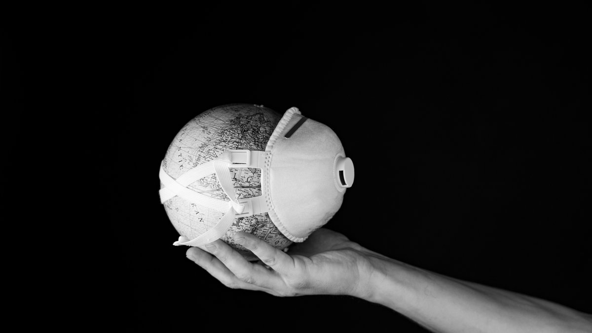 Schwarzweiß-Foto einer Hand, die einen kleinen Globus mit einer Mund-Nasen-Maske hält.