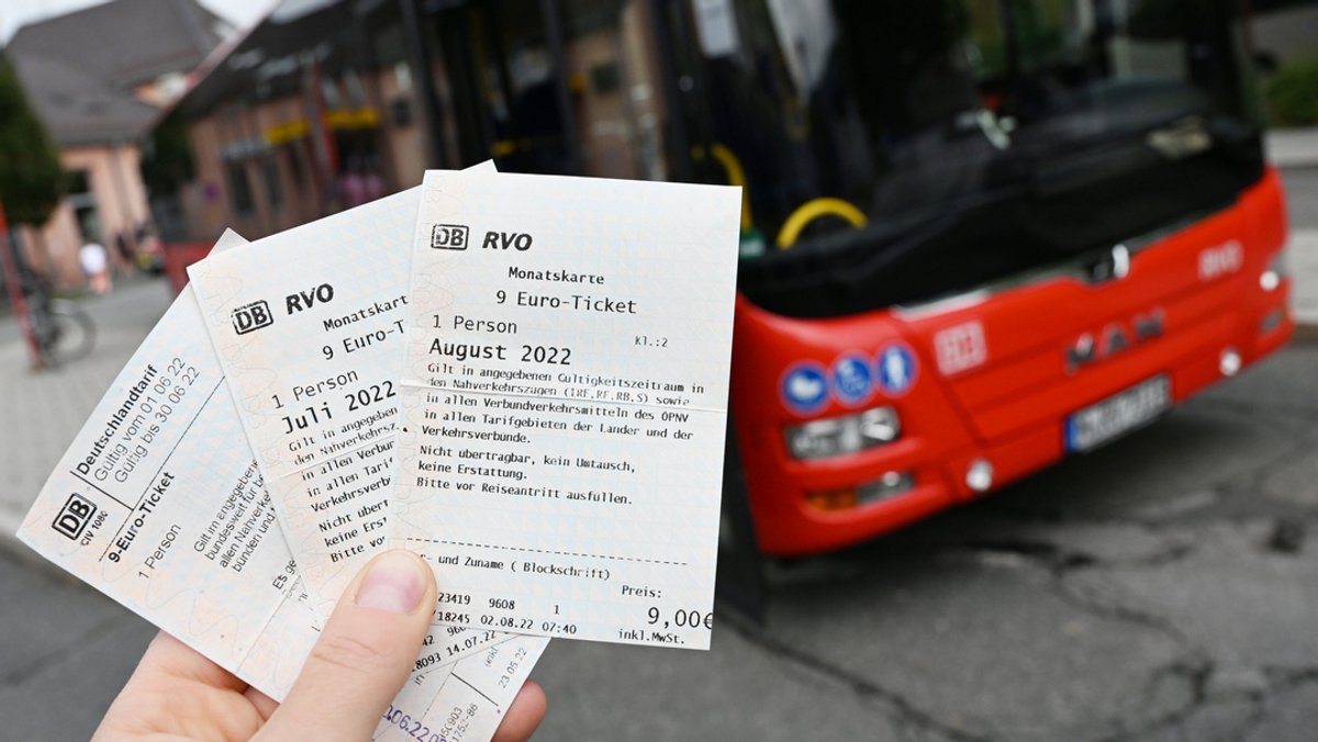 Eine Frau hält drei 9-Euro-Tickets für die Monate Juni, Juli und August vor einem Bus in der Hand.
