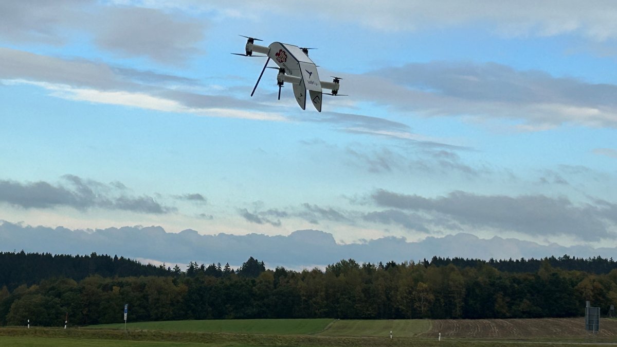 Eine weiße Drohne mit Platz für Medikamente im Inneren fliegt testweise über ein Feld, im Hintergrund ist Wald zu sehen und ein leicht bewölkter Himmel