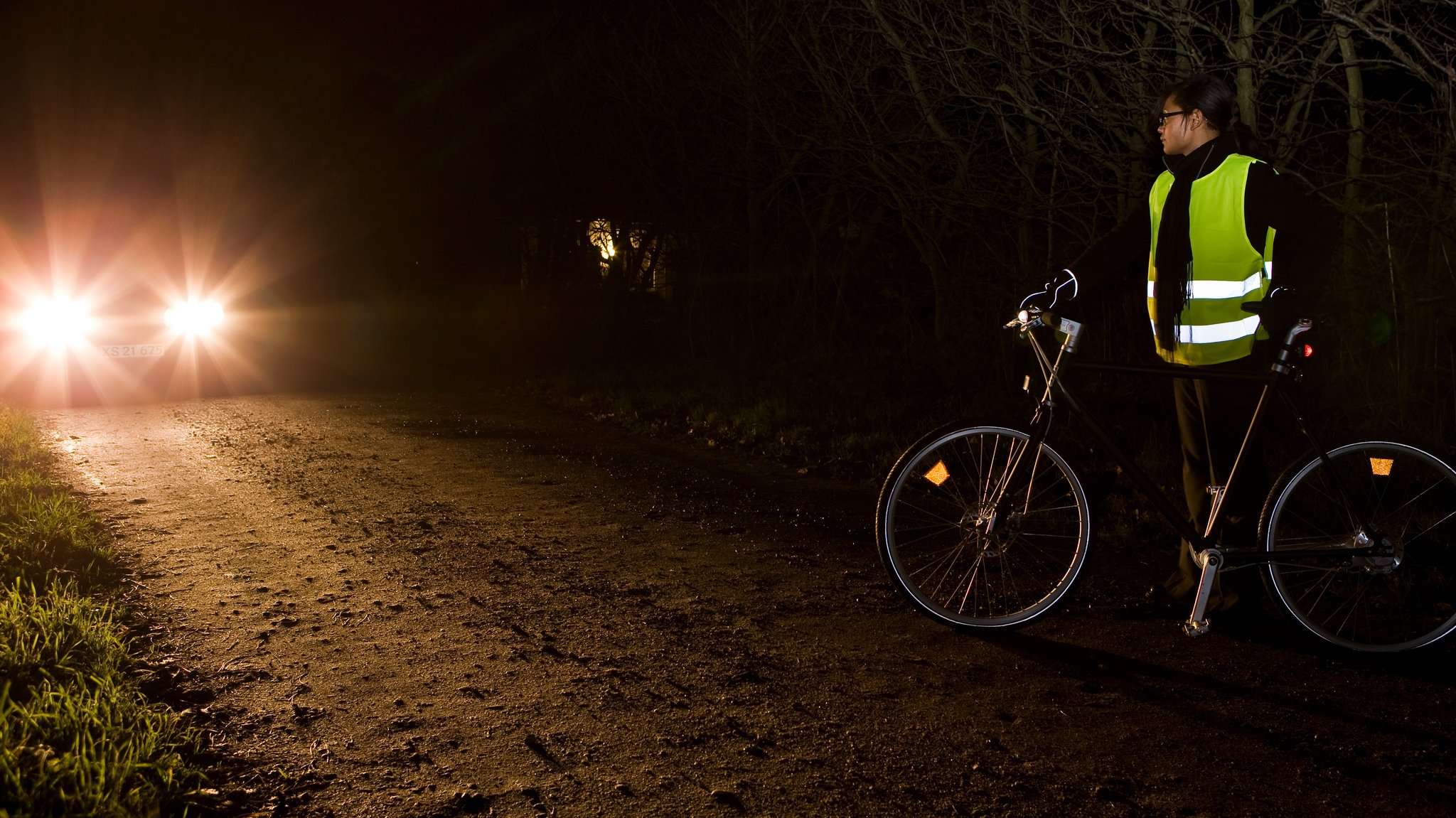 Person mit einem Fahrrad in einer gelben Warnweste wird von Autoscheinwerfern angestrahlt.