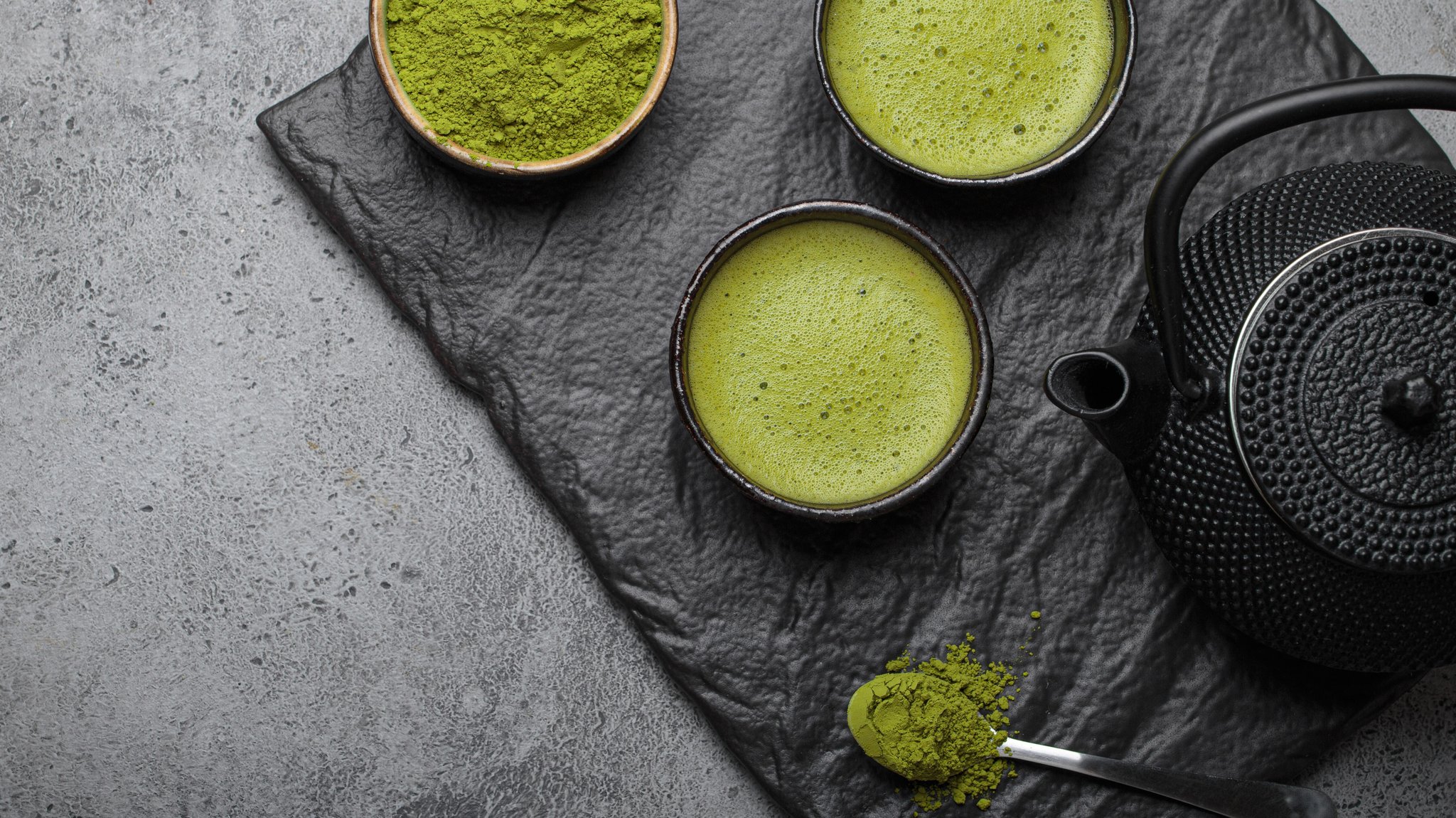 Schwarze Schieferplatte auf der eine schwarze teekanne und zwei Tassen mt Matchatee stehen und eine Schale mit Matchapulver. Ansicht von oben.