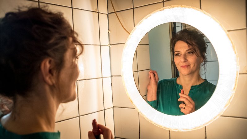Martina Gedeck steht mit einem Lippenstift in der Hand vor einem Badezimmerspiegel
