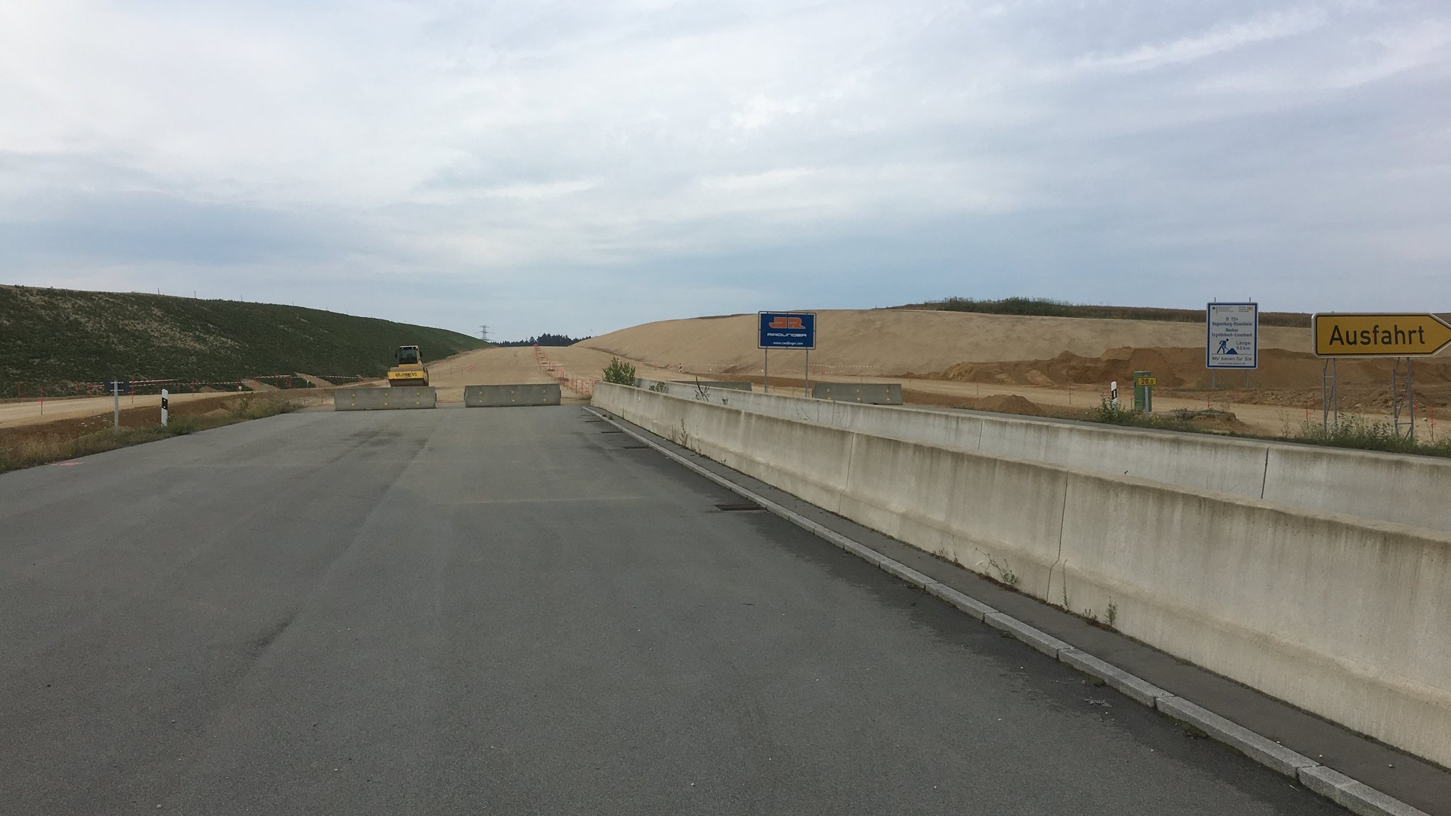 Eine asphaltierte Straße, die sich im Bau befindet und deren vorläufiges Ende mit Betonpoller begrenzt ist.