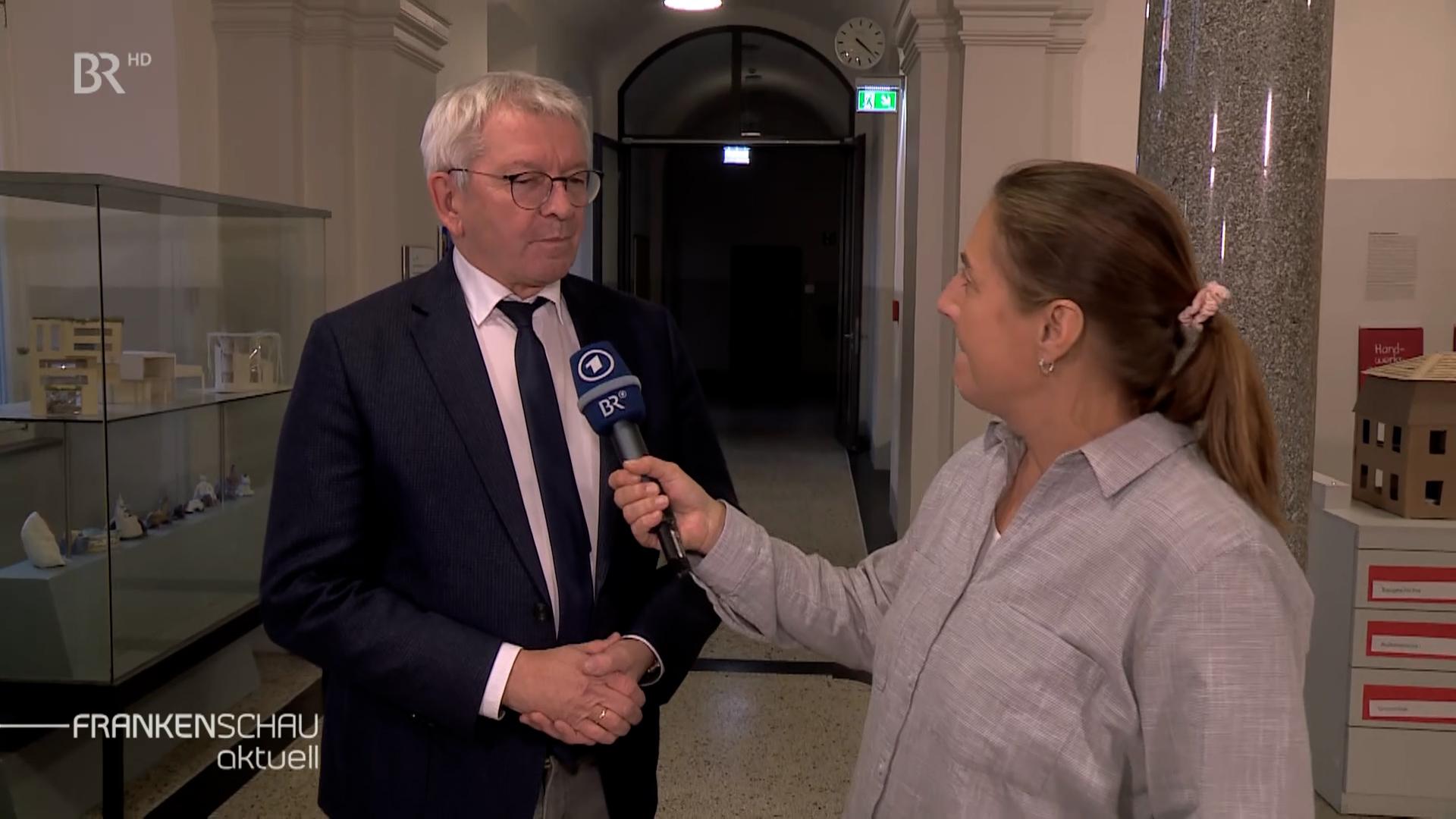 Ein Mann im dunklen Anzug und mit Krawatte wird von einer Reporterin mit einem blauen Mikrophon des Bayerischen Rundfunks interviewt. 