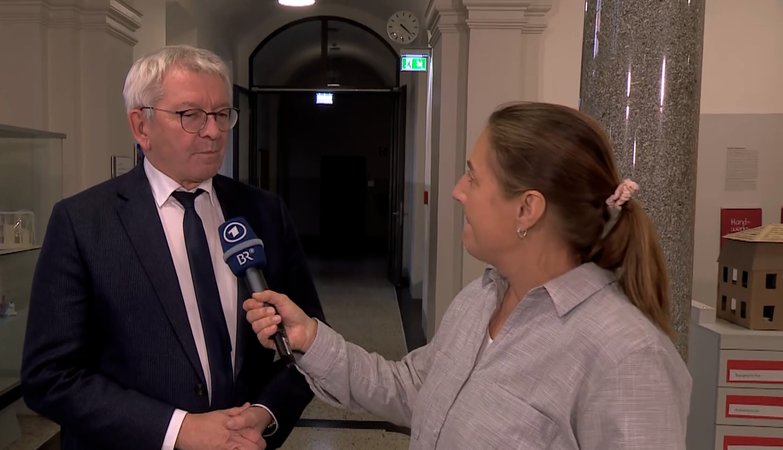 Ein Mann im dunklen Anzug und mit Krawatte wird von einer Reporterin mit einem blauen Mikrophon des Bayerischen Rundfunks interviewt. | Bild: BR Ein Mann im dunklen Anzug und mit Krawatte wird von einer Reporterin mit einem blauen Mikrophon des Bayerischen Rundfunks interviewt.