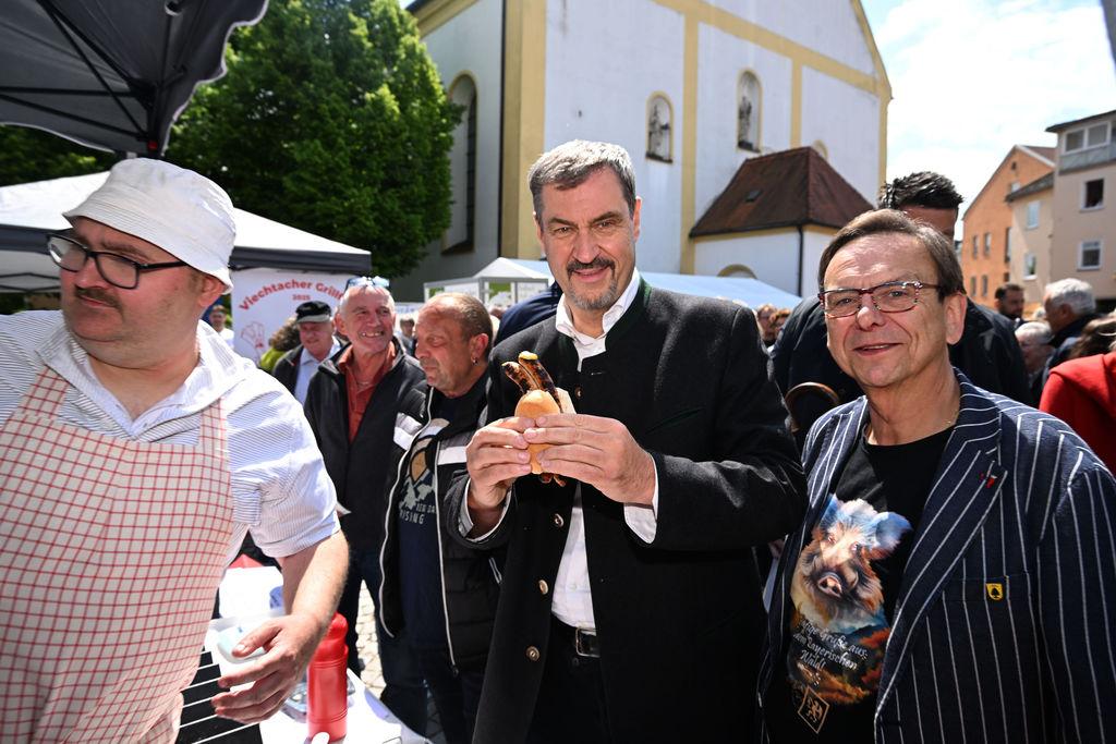 Der bayerische Ministerpräsident Markus Söder hat eine Bratwurstsemmel in der Hand neben Franz Wittmann, dem Veranstalter und Bürgermeister, beim Schweine-Wettgrillen in Viechtach.