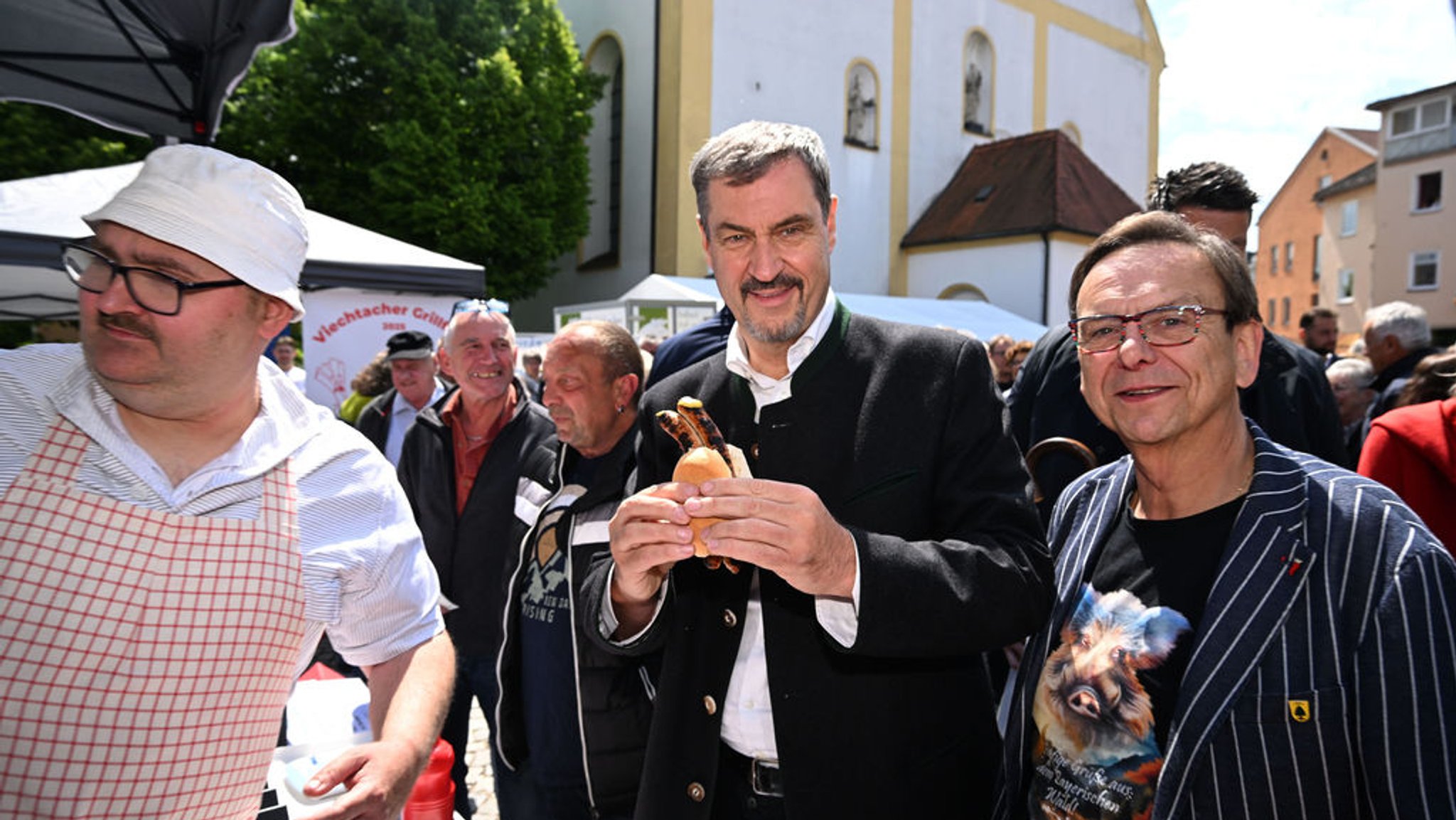 Der bayerische Ministerpräsident Markus Söder hat eine Bratwurstsemmel in der Hand neben Franz Wittmann, dem Veranstalter und Bürgermeister, beim Schweine-Wettgrillen in Viechtach.