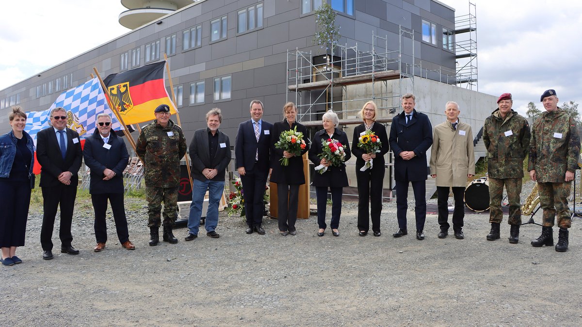 Mehrere Persönlichkeiten aus Politik und Wirtschaft stehen, teils mit Blumensträußen, für ein Gruppenbild vor dem neuen Gebäude für technische Aufklärung der Bundeswehr. 