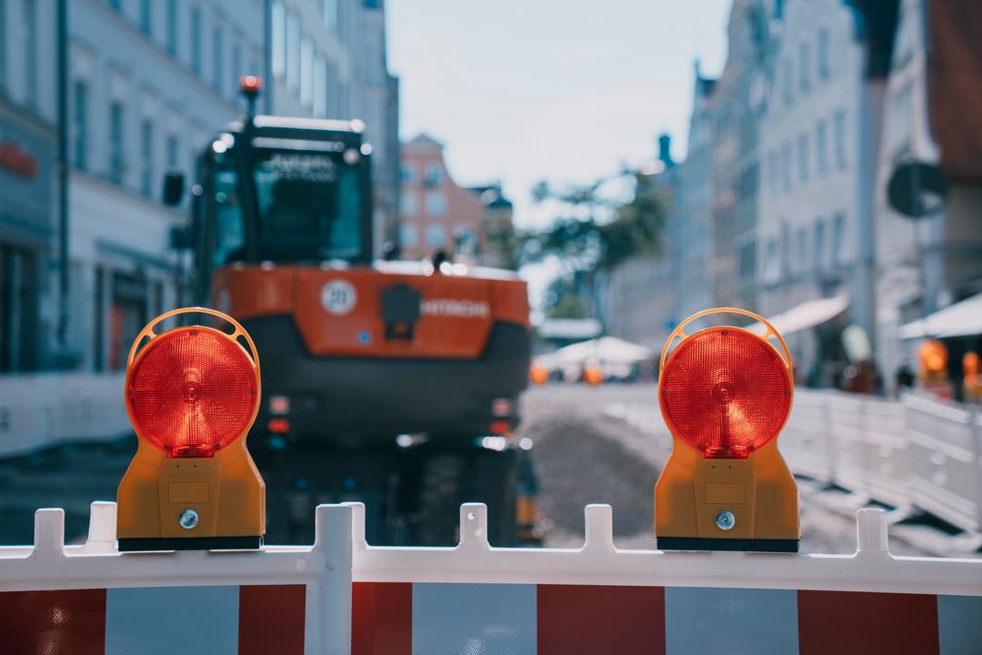 Zwei rote Baustellenleuchten auf einem Absperrzaun in der Fußgängerzone. Dahinter in der Unschärfe ein geparkter Bagger.