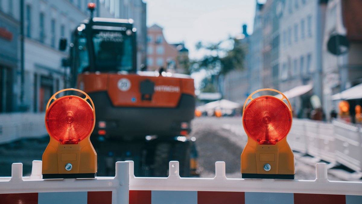 Zwei rote Baustellenleuchten auf einem Absperrzaun in der Fußgängerzone. Dahinter in der Unschärfe ein geparkter Bagger.