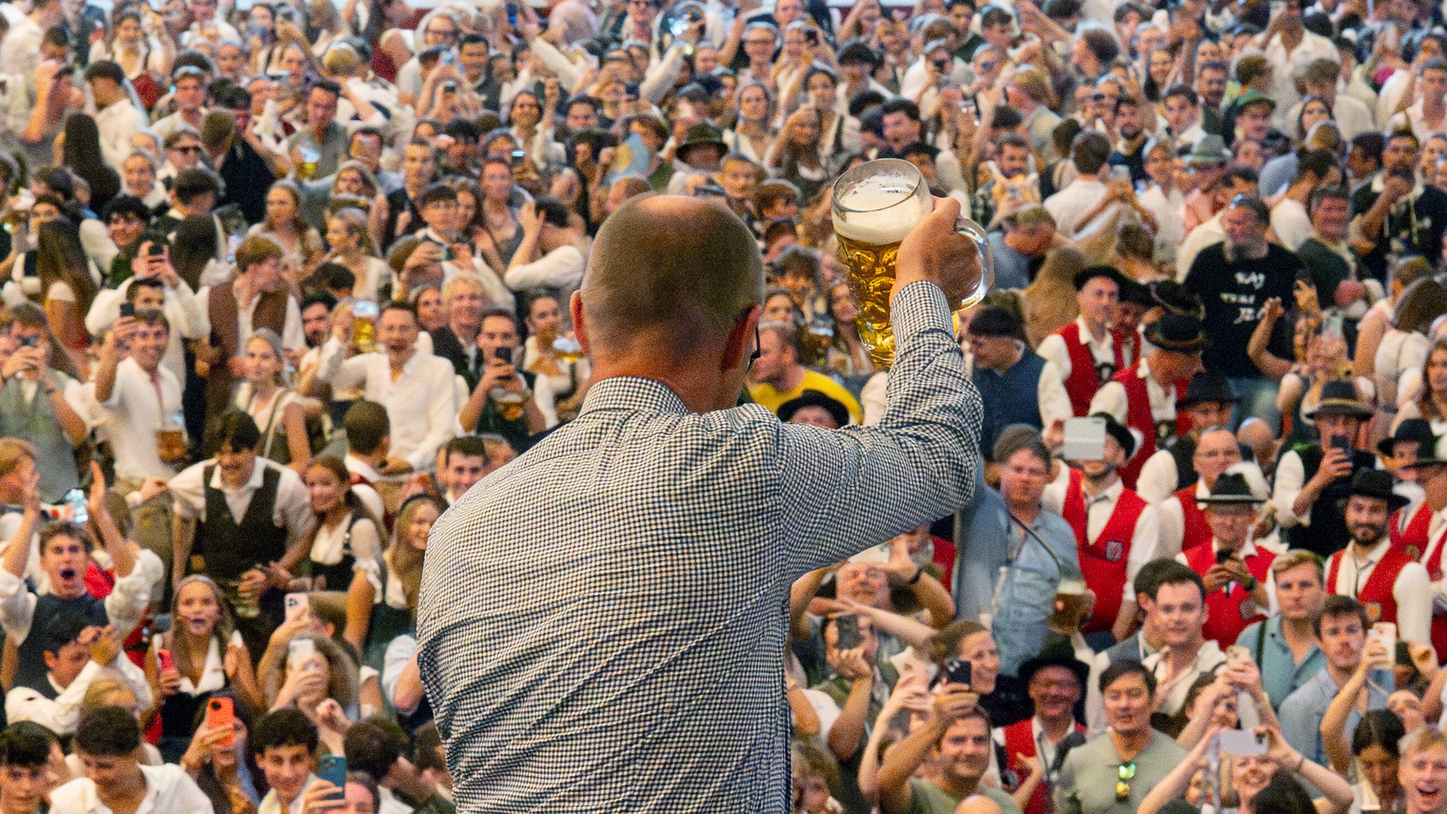 Bundeskanzler Merz im karierten Hemd in der Rückenansicht prostet vom Balkon des Festzelts der Menge unten zu. Merz hat dem Oktoberfest einen Besuch abgestattet.