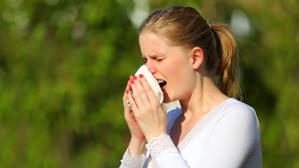 Frau in weißem T-Shirt niest in ein Taschentuch. Im Hintergrund verschwommen grüne Natur.