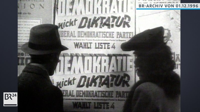 Ein Mann links und eine Frau rechts stehen vor einer Plakatwand. Auf dem Plakat steht "Demokratie nicht Diktatur". Liebarl Demokratische Partei.
