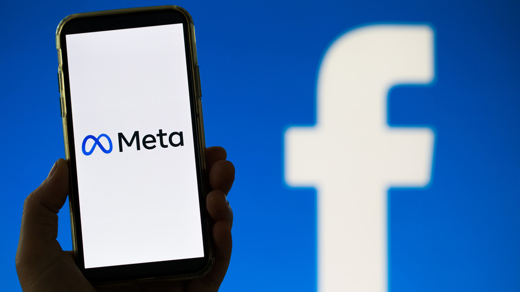 Smartphonebildschirm, auf dem das Meta-Logo zu sehen ist. Im Hintergrund ein großes Facebook-Logo.