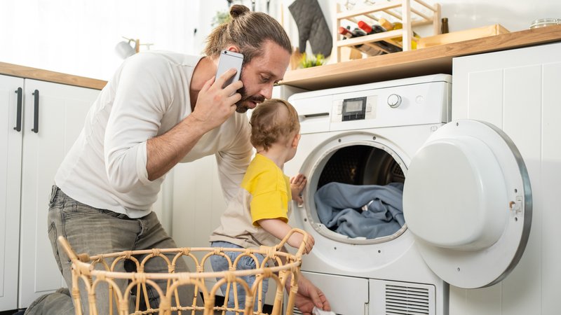 Ein junger Vater telefoniert mit dem Handy, während er eine Waschmaschine einräumt und ein Kleinkind betreut. | Bild: stock.adobe.com/Kawee Ein junger Vater telefoniert mit dem Handy, während er eine Waschmaschine einräumt und ein Kleinkind betreut.