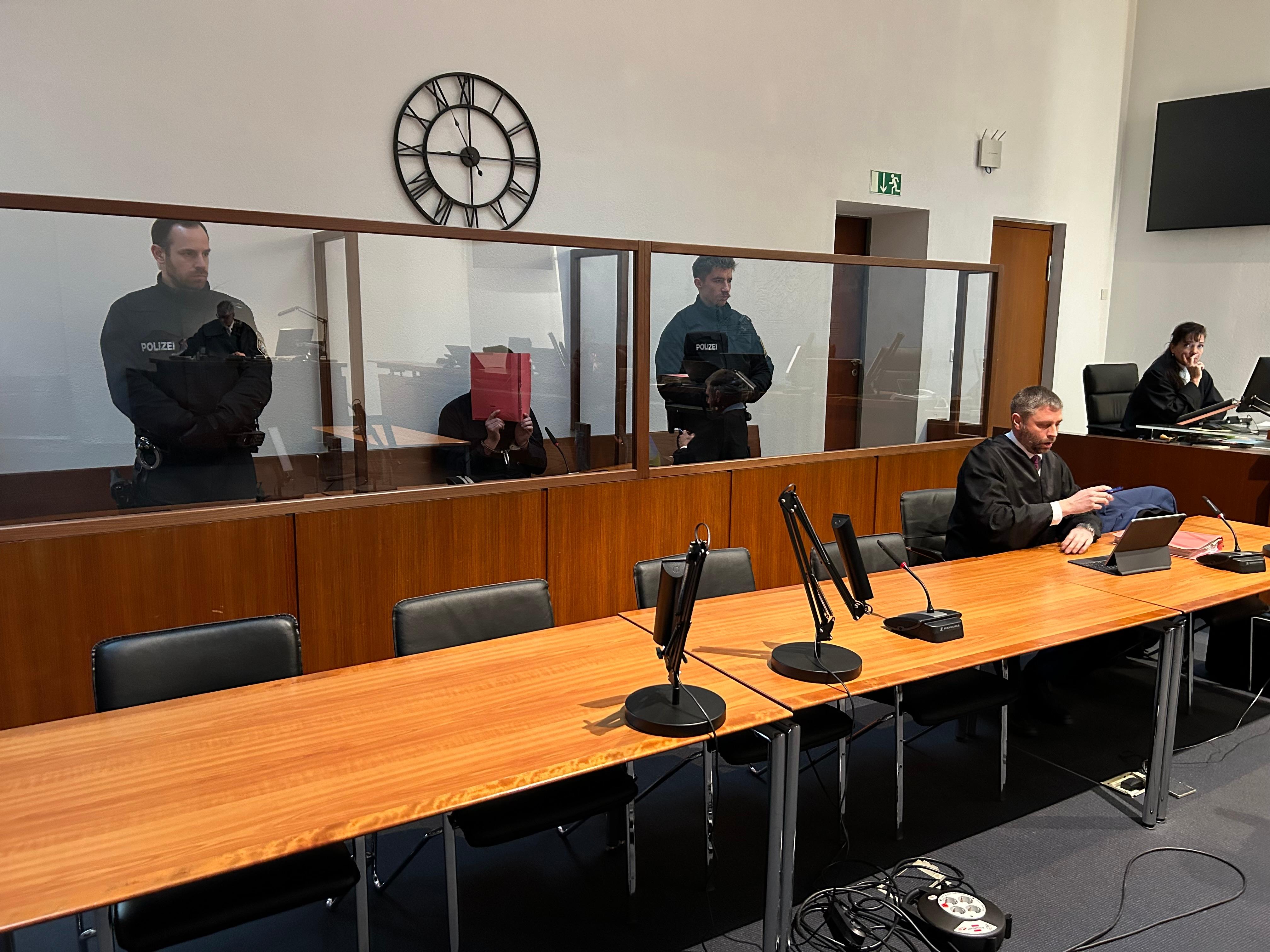 Ein Angeklagter sitzt auf der Anklagebank und hält sich einen Aktenordner vors Gesicht. Neben ihm stehen zwei Polizisten. 