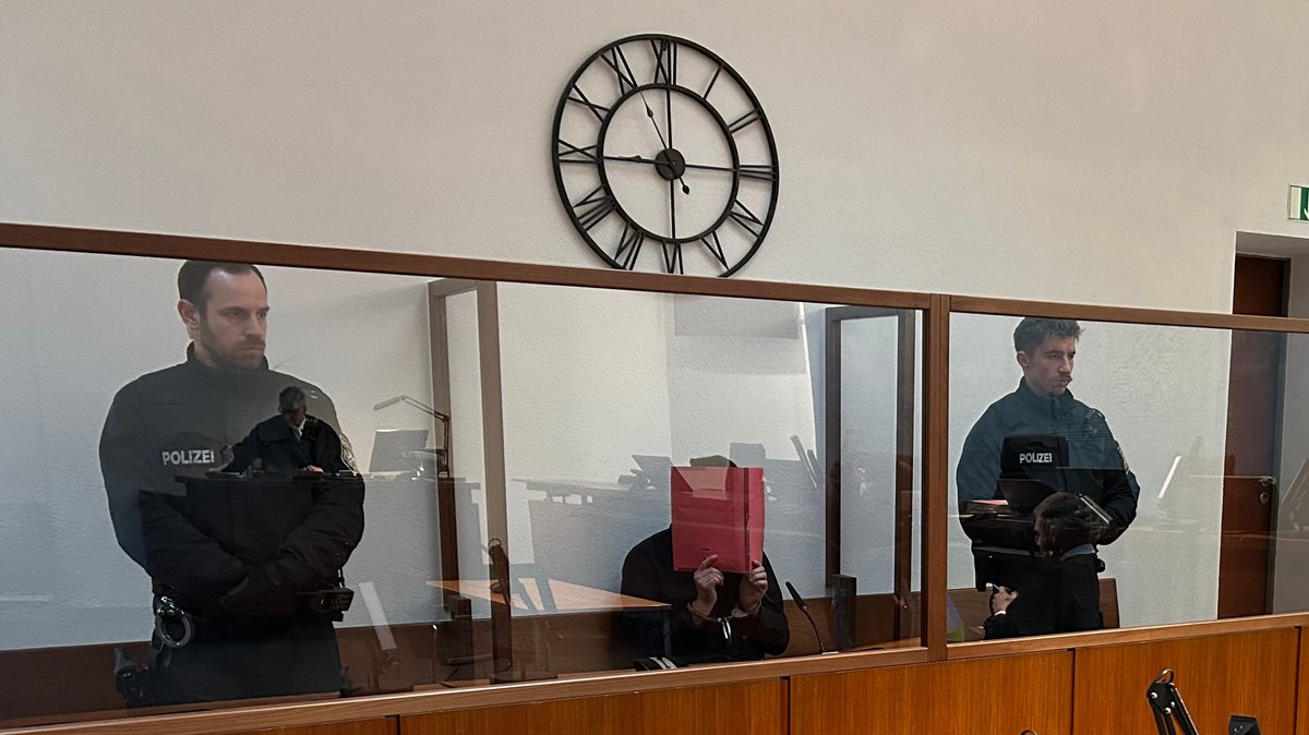 Ein Angeklagter sitzt auf der Anklagebank und hält sich einen Aktenordner vors Gesicht. Neben ihm stehen zwei Polizisten. 