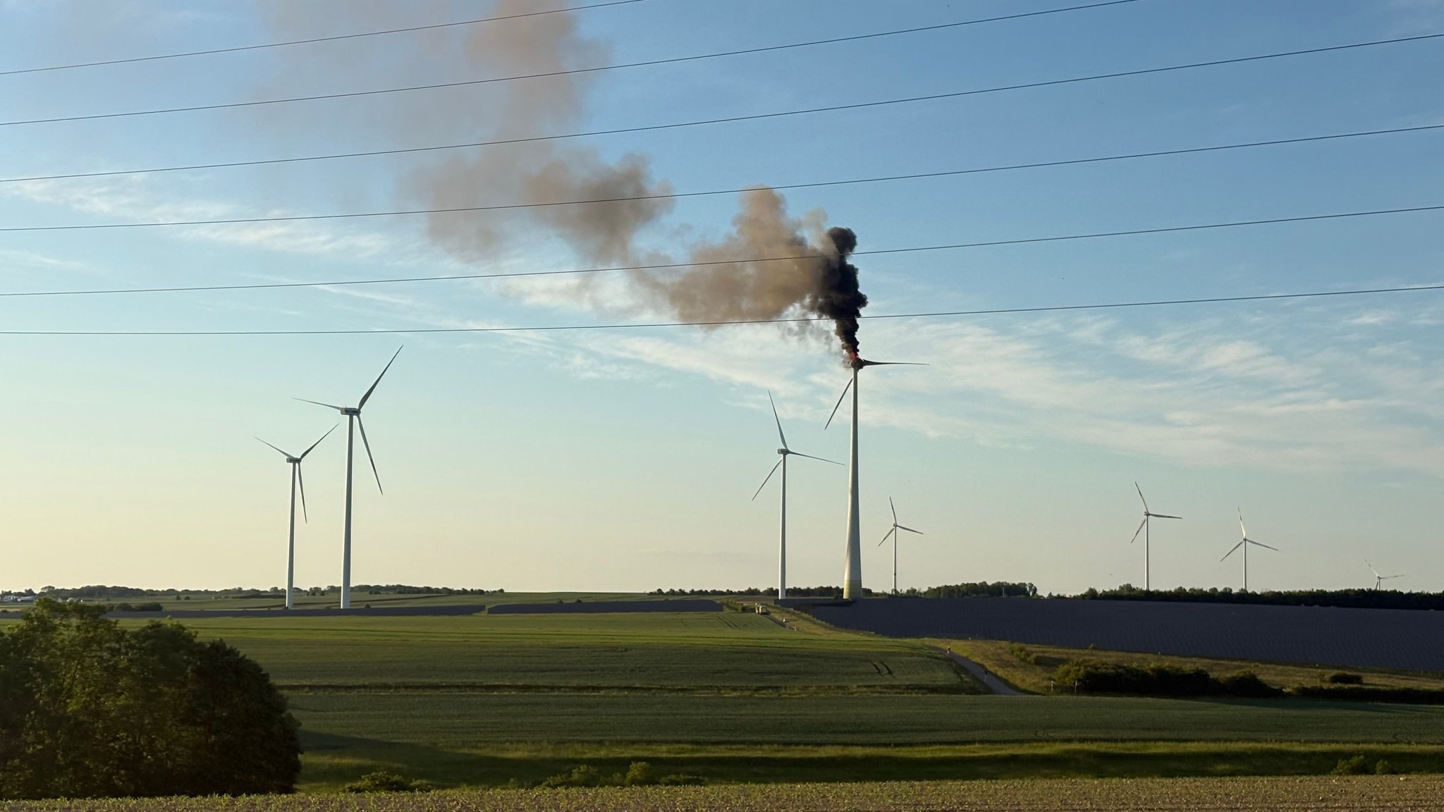 Mehrere Windräder auf einem freien Feld, von dem eines brennt. Schwarzer Rauch steigt von ihm den Himmel hoch.