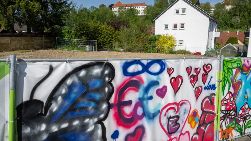 Ein Bauzaun ist mit Graffiti dekoriert und versperrt den Blick auf ein leeres Grundstück.