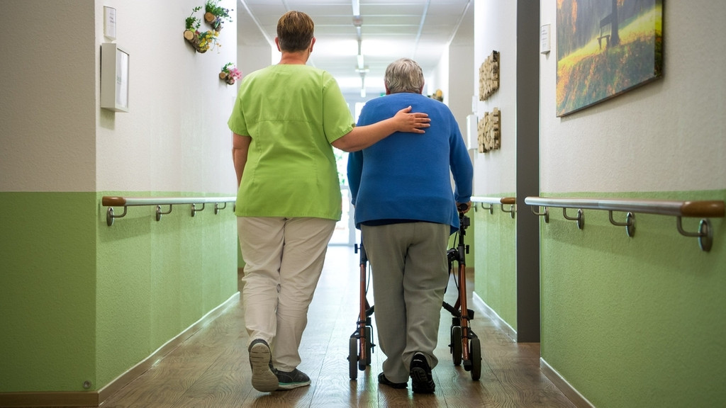 Eine Altenpflegerin begleitet eine Seniorin, die an einem Rollator geht, über den Flur einer Pflegeeinrichtung (Symbolbild)