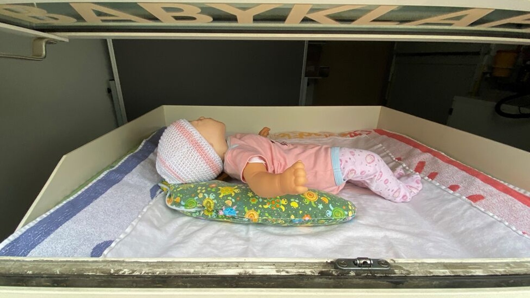 05.01.2021, Regensburg - Zu Demonstrationszwecken liegt eine Puppe in der Babyklappe der Klinik St. Hedwig in Regensburg. Die Babyklappe befindet sich in einem geschützten Bereich, der von außen zugänglich ist. In der Klappe befindet sich ein Bett, in das das Baby hineingelegt werden kann. Durch das Hineinlegen wird ein Sensor aktiviert, der anschließend mit einer 30-sekündigen Zeitverzögerung beim Klinikpersonal ein Alarm ertönen lässt. | Bild: BR/Zara Kroiß 05.01.2021, Regensburg - Zu Demonstrationszwecken liegt eine Puppe in der Babyklappe der Klinik St. Hedwig in Regensburg. Die Babyklappe befindet sich in einem geschützten Bereich, der von außen zugänglich ist. In der Klappe befindet sich ein Bett, in das das Baby hineingelegt werden kann. Durch das Hineinlegen wird ein Sensor aktiviert, der anschließend mit einer 30-sekündigen Zeitverzögerung beim Klinikpersonal ein Alarm ertönen lässt.