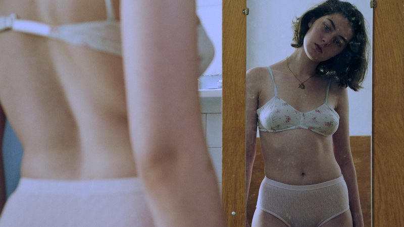 Junge Frau steht in Unterwäsche vor dem Spiegel und betrachtet ihren Körper.
