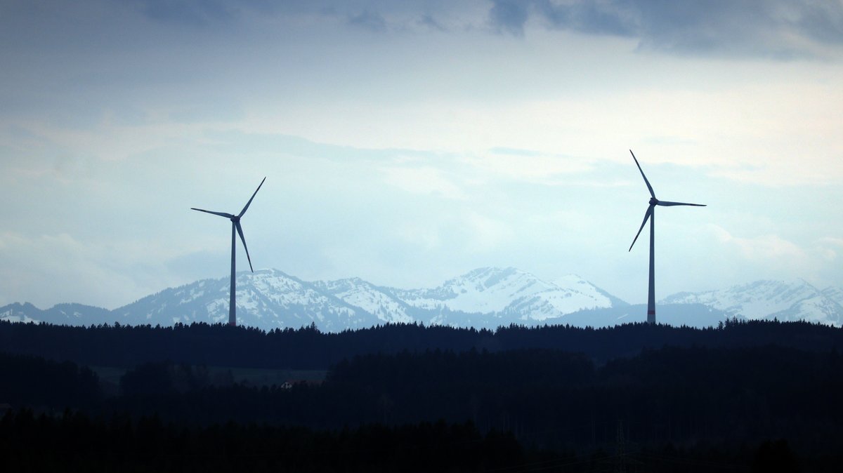 Zwei Windräder stehen vor den schneebedeckten Alpen.