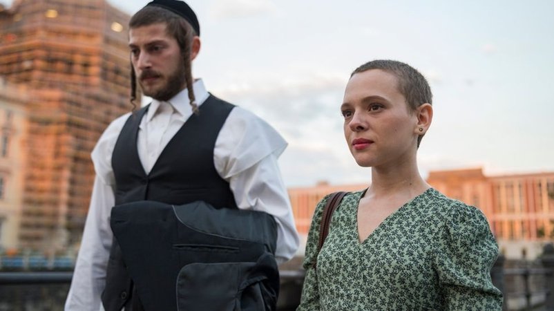 Ein Mann in jüdisch-orthodoxer Kleidung und eine Frau mit Pixie-Frisur laufen auf der Straße: Szene aus "Unorthodox"