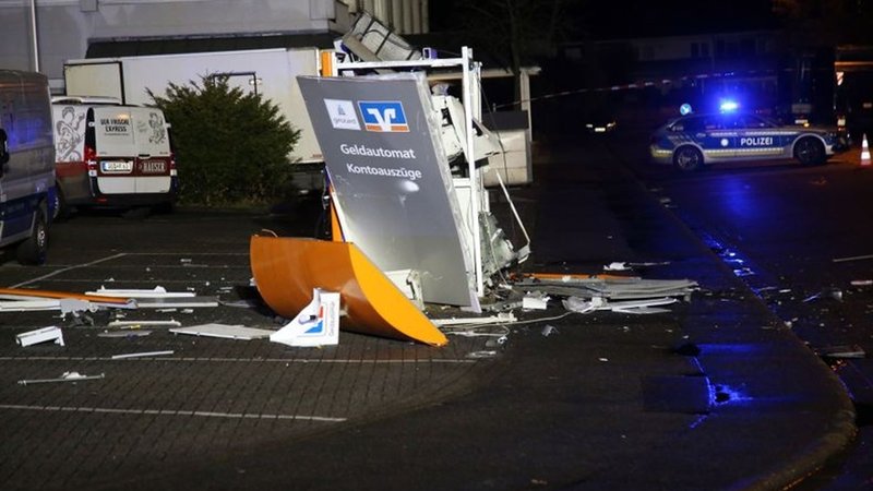 Ein gesprengter Geldautomat im Dunkeln, drumherum Trümmerteile. Im Hintergrund ein Polizeiauto mit Blaulicht. 
