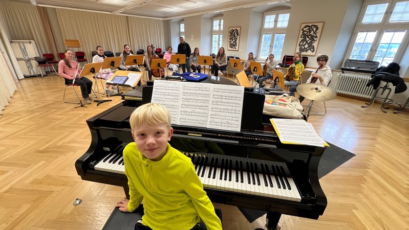 Ein Junge sitzt vor einem Flügel, im Hintergrund ein Schulorchester. | Bild: BR / Rupert Waldmüller Ein Junge sitzt vor einem Flügel, im Hintergrund ein Schulorchester.